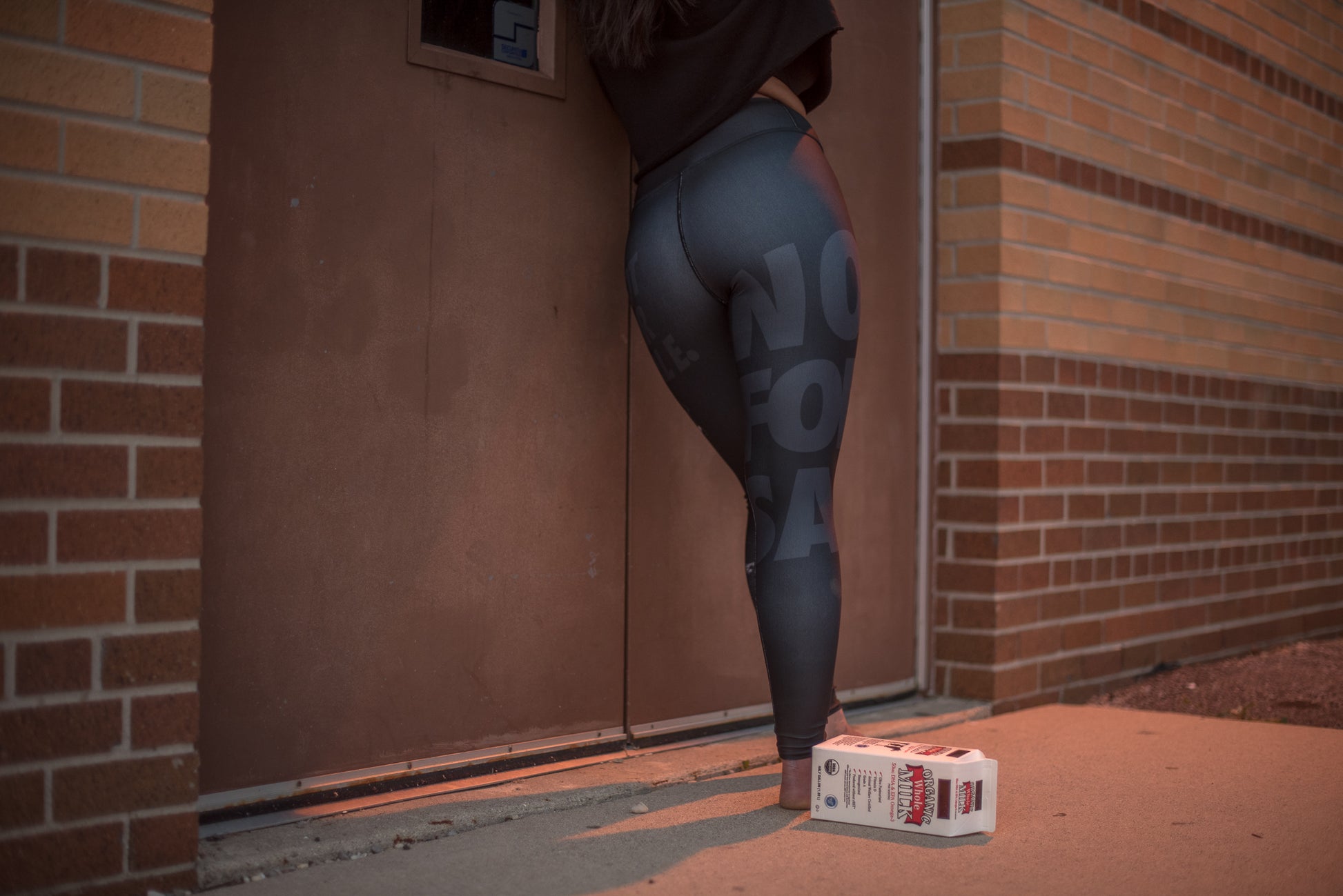 Person wearing leggings with text standing next to a box of beer against a brick wall.